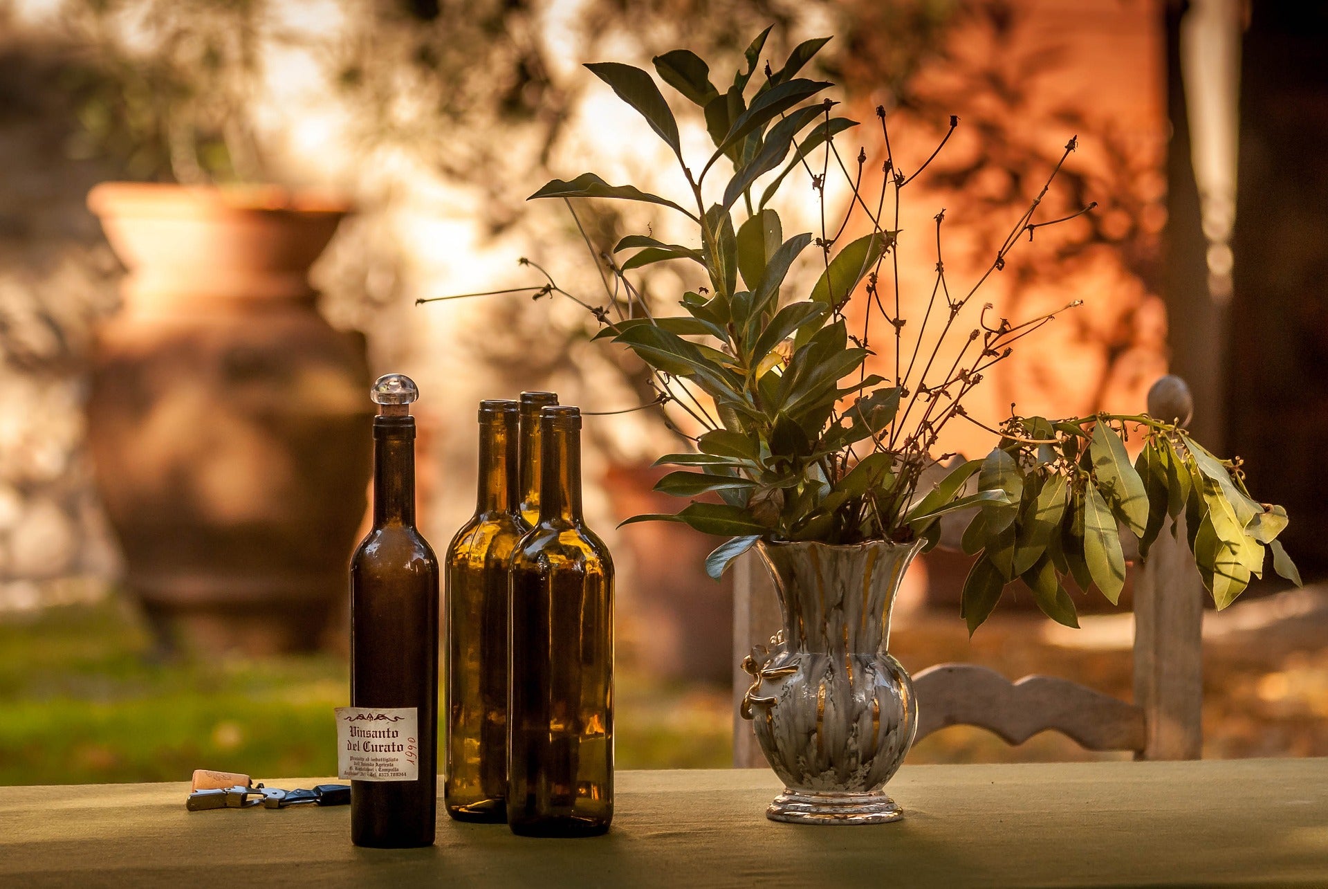 Fire vinflasker og en dekorativ vase med grønne grene på et bord i udendørs omgivelser.