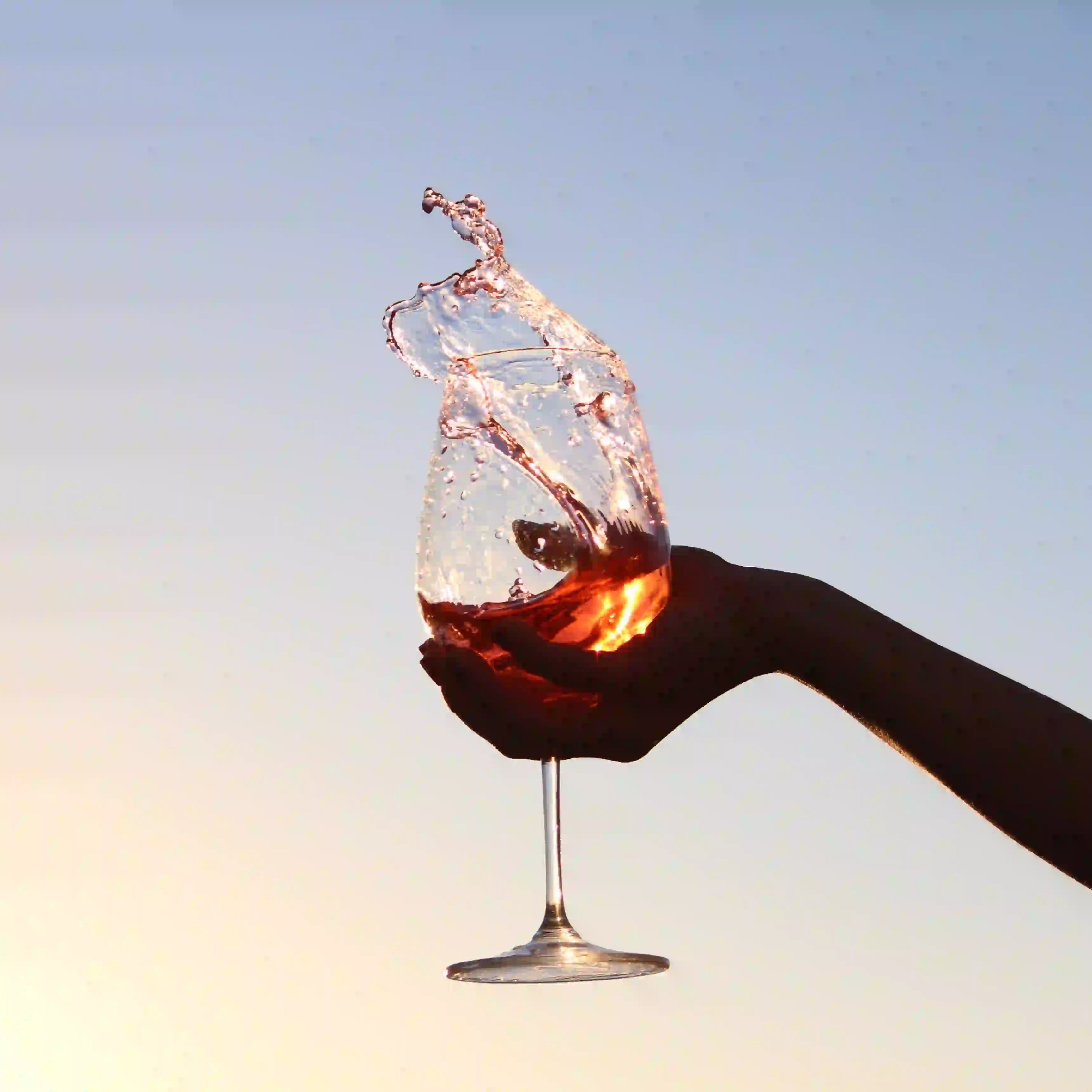 Hånd, der holder et glas rosévin mod himlen, mens vinen sprøjter op i luften i solnedgangens varme lys.
