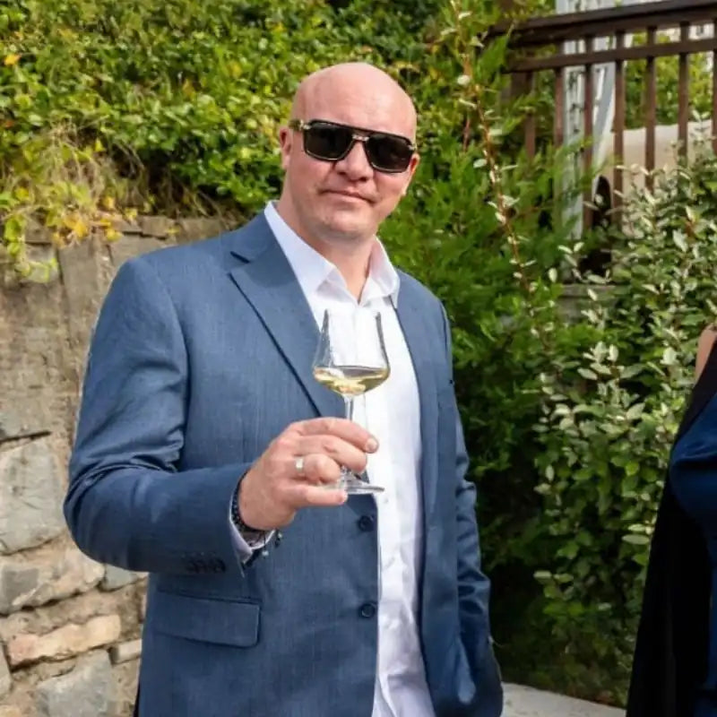 Ejer af Bottle Butler i lyseblå habitjakke, hvid skjorte og solbriller står udendørs med et glas hvidvin i hånden og smiler mod kameraet.