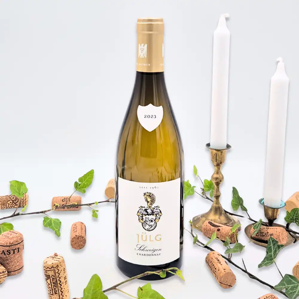 Schweigen chardonnay omgivet af vinkorker, grønne blade og to hvide lysestager på hvid baggrund.