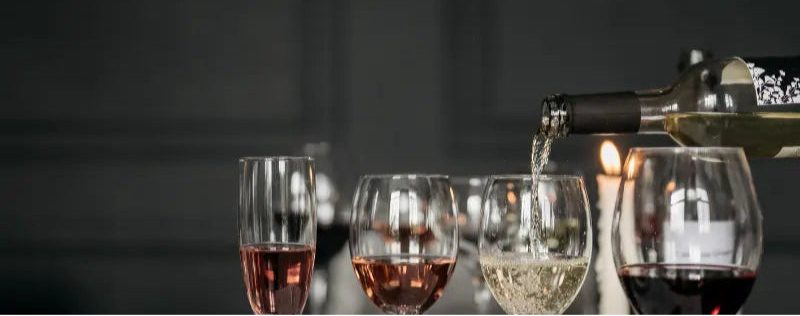 Række af vinglas med rødvin, rosé og hvidvin, hvor der skænkes hvidvin i et glas på et bord med tændte stearinlys i baggrunden.