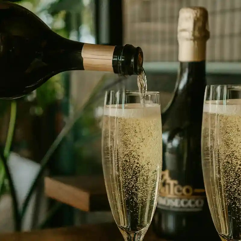 Flaske mousserende vin hældes op i et glas med fine bobler, mens en flaske prosecco står i baggrunden.