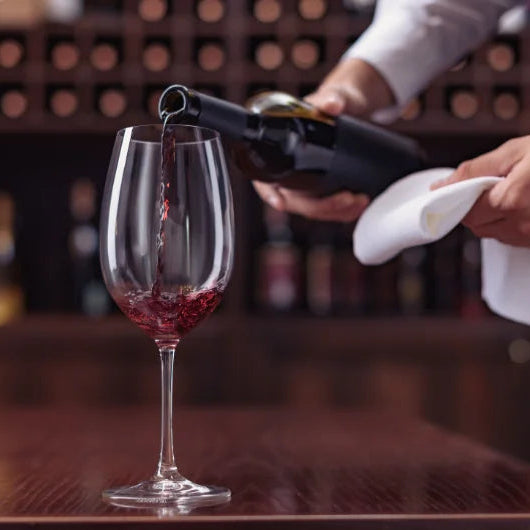 Butler hælder rødvin i et vinglas på en restaurant eller vinbar med vinreol i baggrunden.