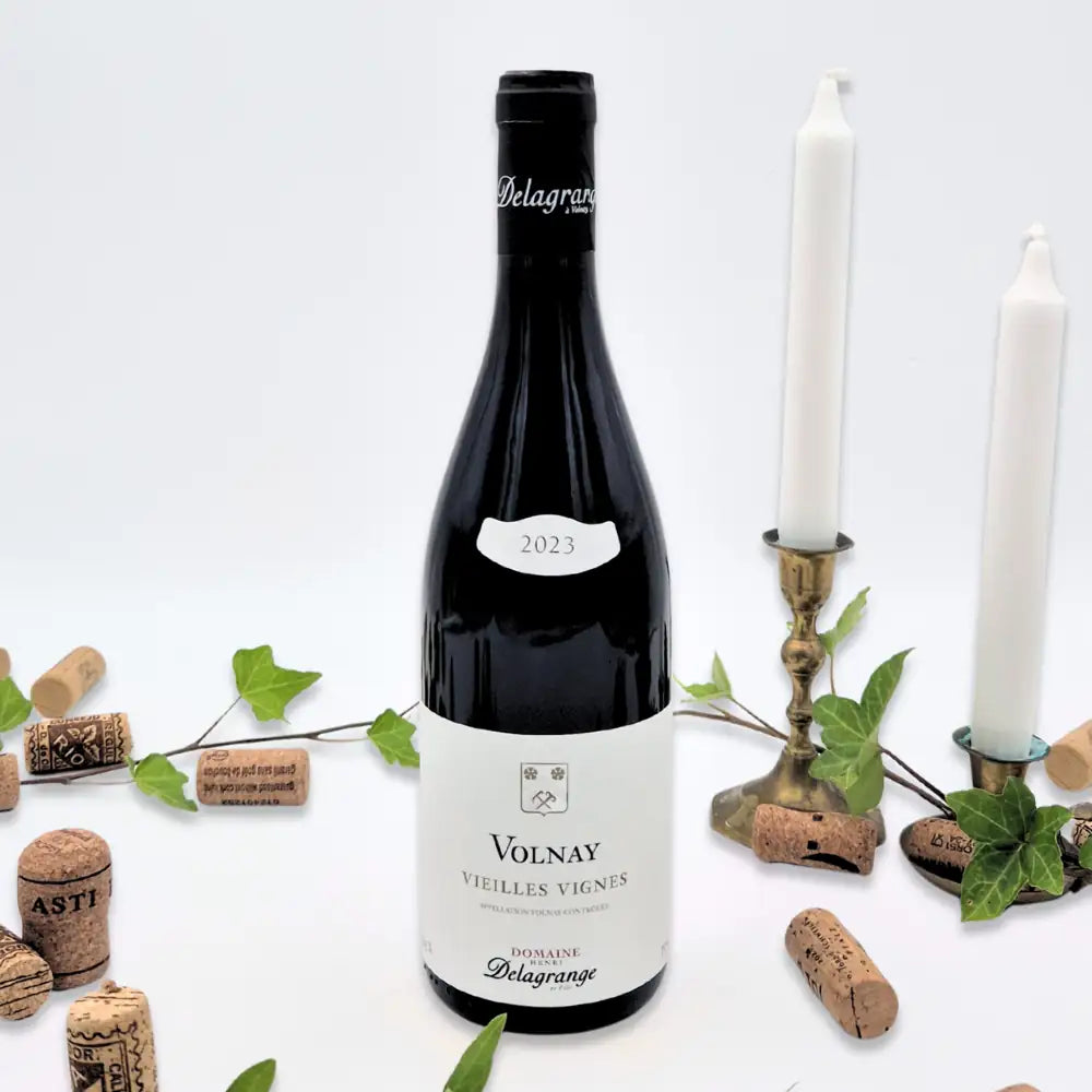 Volnay Vielles Vignes omgivet af vinkorker, grønne blade og to hvide lysestager på hvid baggrund.