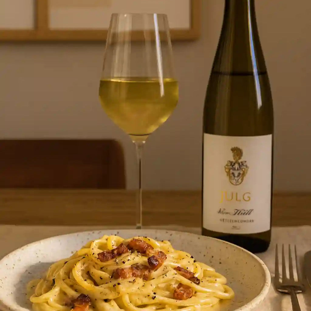 Tallerken med cremet pasta carbonara serveret med et glas hvidvin og en flaske Jülg Riesling von Kalk på et spisebord i varm belysning.
