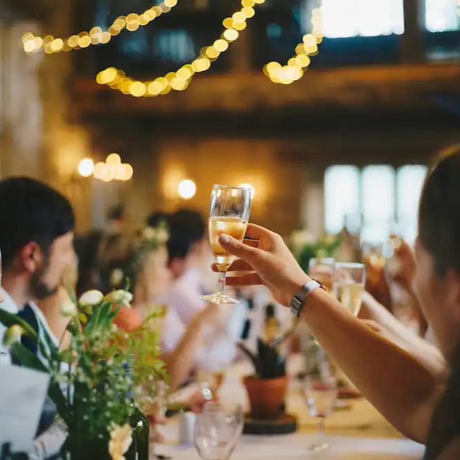 Festligt selskab med gæster, der skåler med champagneglas i et smukt oplyst lokale dekoreret med blomster og lyskæder.