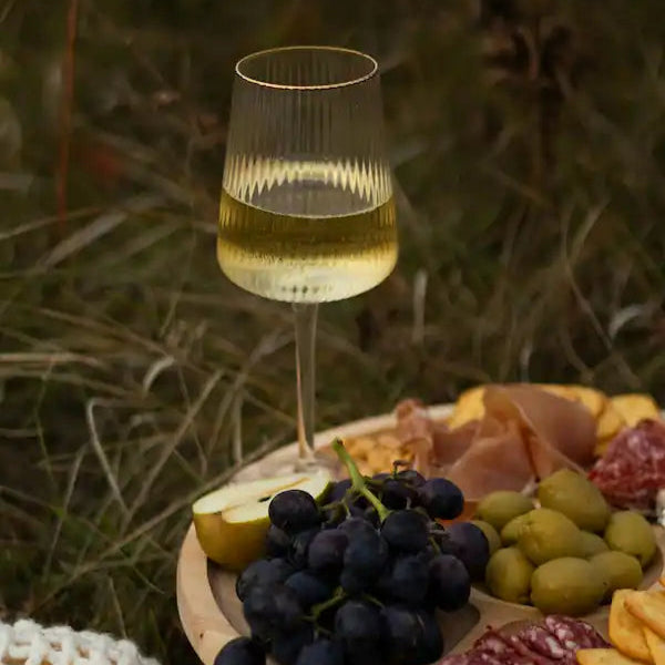 Glas hvidvin placeret på et træfad med druer, oliven, skinke og ost i græsset under en udendørs picnic.