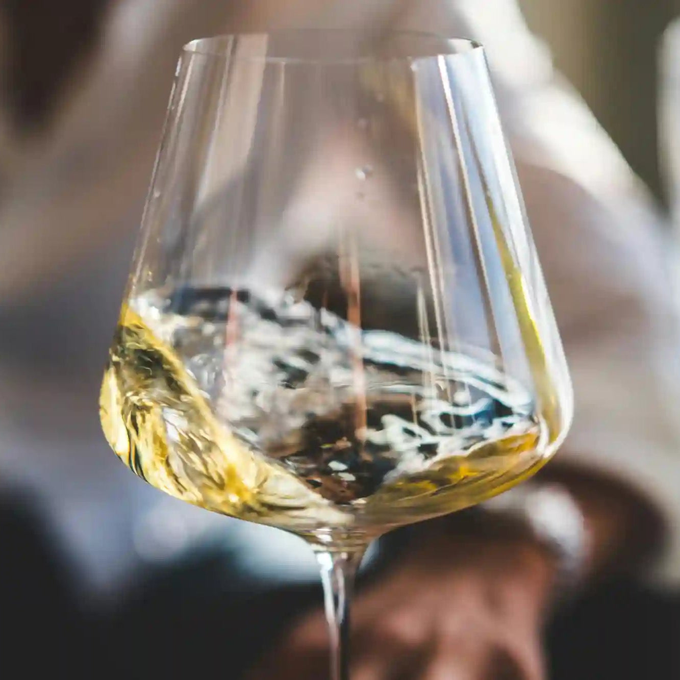 Nærbillede af et vinglas med hvidvin, der slynges i glasset, med en person i baggrunden ude af fokus.