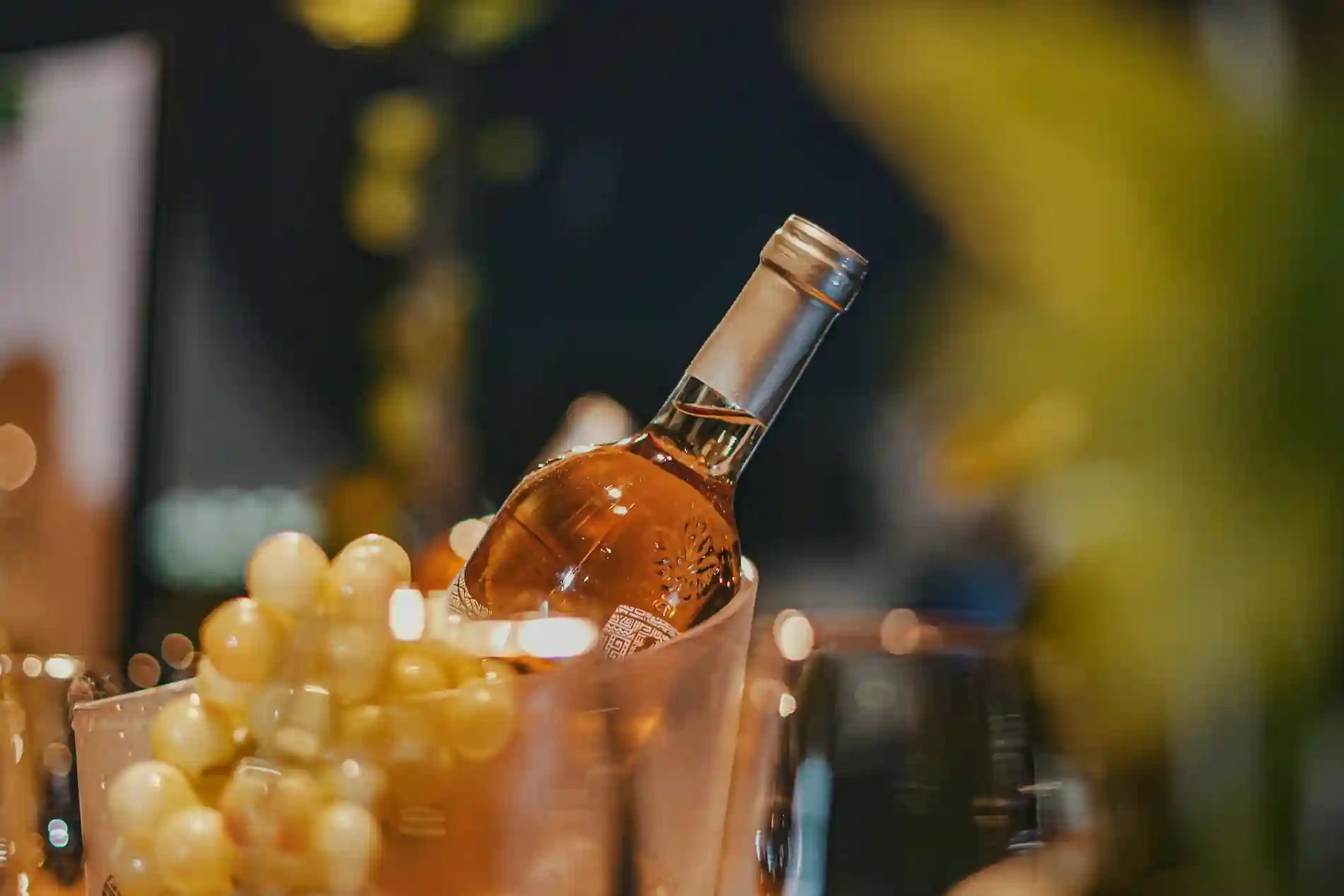 Flaske rosévin placeret i en vinkøler med is, omgivet af vindruer og varm belysning i baggrunden.