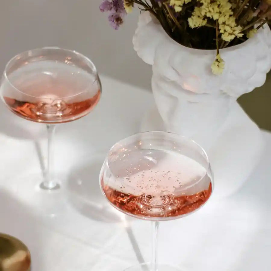 To glas med rosévin står på et hvidt bord ved siden af en hvid vase med tørrede blomster.