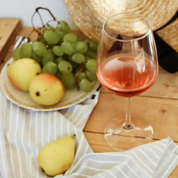 Glas med rosévin på et træbord sammen med grønne druer, pærer og æbler, med en stråhat i baggrunden.