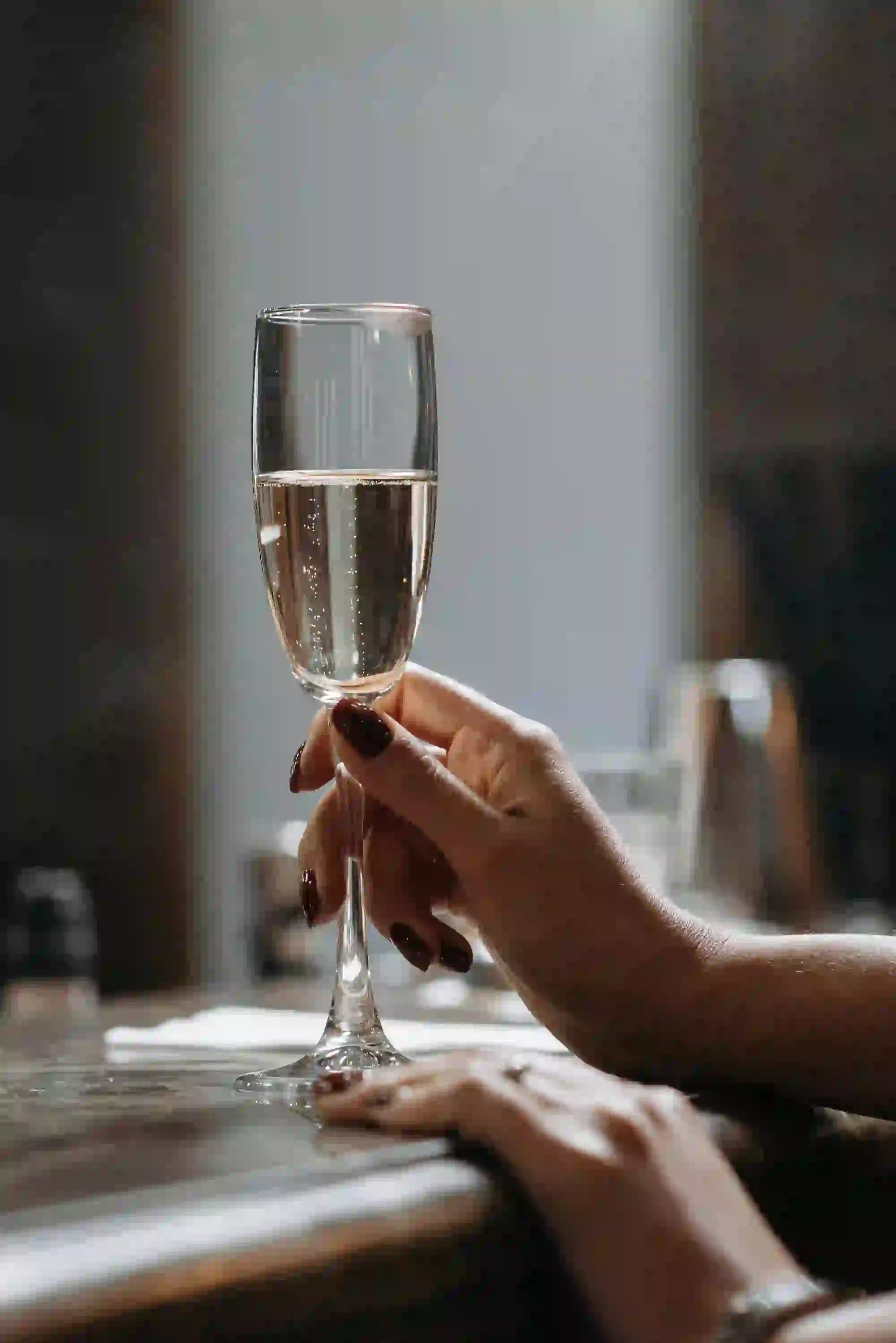 Hånd med mørkerøde negle, der holder et glas mousserende vin ved et bord i afdæmpet belysning.