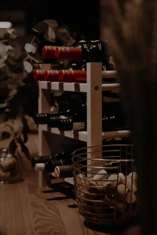 Vinreol i lyst træ fyldt med vinflasker, omgivet af grønne planter og en kurv med vinpropper i forgrunden.